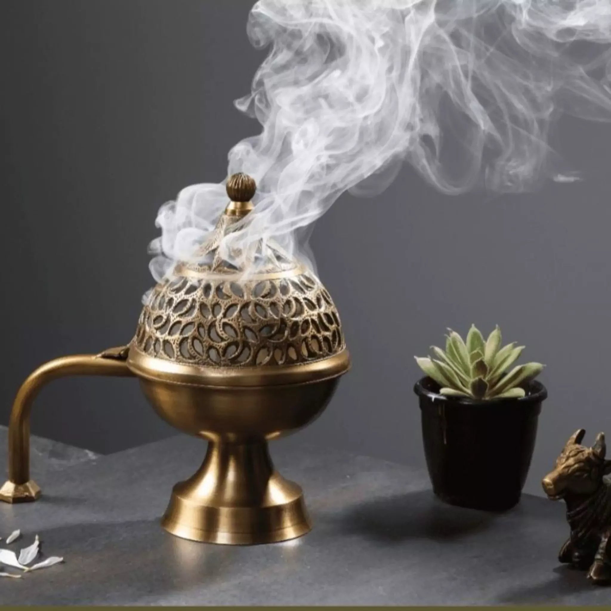Bronze incense burner with smoke rising, next to a small potted plant and figurine on a dark surface.